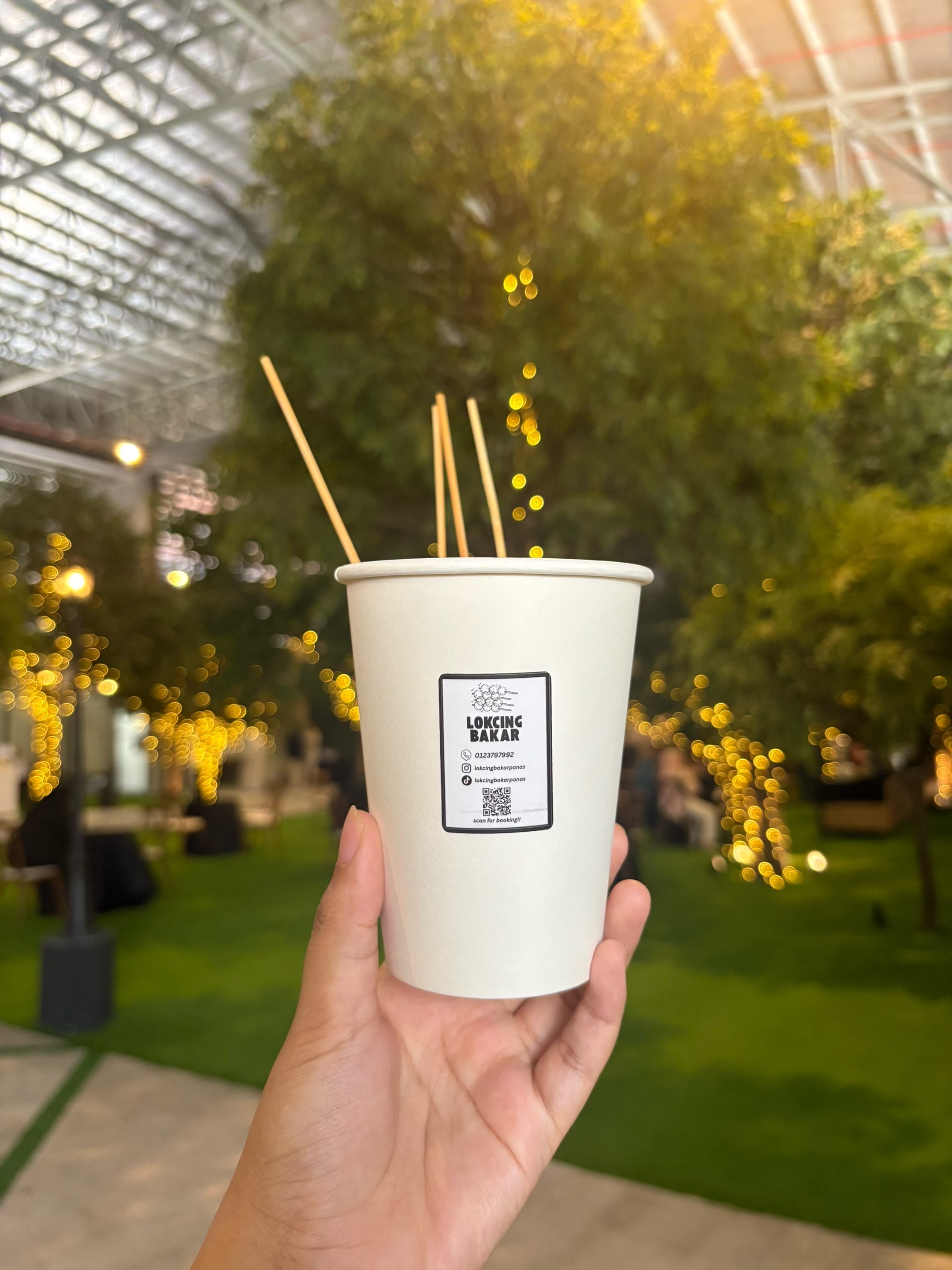 Cup Lokcing Bakar dipegang di kawasan majlis dengan lampu hiasan
